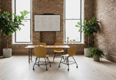 Aarhus Eiffel Chair V2 by Quell Design in warm ginger orange placed around a meeting table in a bright contemporary office with exposed red brick walls, large industrial-style windows, wooden floors, and lush green plants. These Scandinavian-inspired ergonomic chairs feature smooth moulded polypropylene seats, black swivel wheel bases, and under-seat storage, offering comfort, mobility, and minimalist design for collaborative workspaces.
