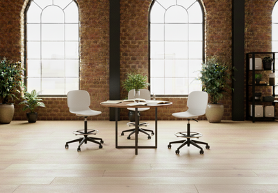 Aarhus Swivel Counter Stools by Quell Design in white arranged around a round wooden table in a modern industrial-style room with exposed brick walls, tall windows, and warm wooden floors. These Scandinavian-inspired adjustable stools feature smooth moulded polypropylene seats, black five-star swivel bases, and integrated footrests, combining ergonomic comfort and minimalist design for contemporary office or café spaces.
