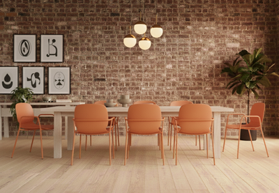 Aarhus Bistro Arm Chairs by Quell Design in coral arranged around a long natural wooden table in a modern industrial interior with exposed red brick walls and light white oak flooring. The Scandinavian-style chairs feature ergonomic moulded polypropylene seats with integrated armrests and slim black powder-coated steel legs, creating a warm and collaborative workspace or café atmosphere.