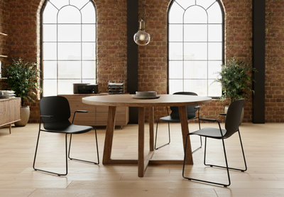 Aarhus Bistro Carver Chairs by Quell Design in black arranged around a round wooden table in a contemporary interior with exposed red brick walls, large industrial-style windows, and warm wooden furniture. These Scandinavian-inspired chairs feature ergonomic moulded polypropylene seats with integrated armrests and slim black powder-coated steel legs, creating a sophisticated and collaborative workspace or café environment.
