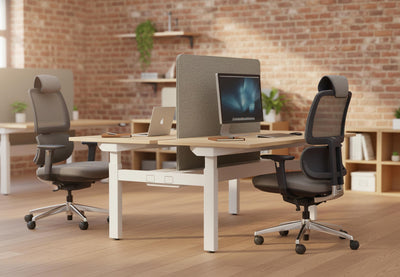 Anka Adaptive Desk Chair in black shown in a modern office interior, featuring two chairs positioned at work desks within a space with exposed brick walls, wooden flooring, natural light and greenery, highlighting contemporary ergonomic office seating and professional workplace design.