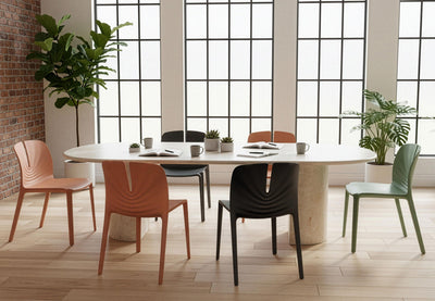 Six Emi Stackable Office Seating chairs in morandi green, coral and black arranged around a large marble collaboration table in an office setting with exposed brick walls, wooden flooring, tall windows and greenery, showcasing modern stackable workspace seating.