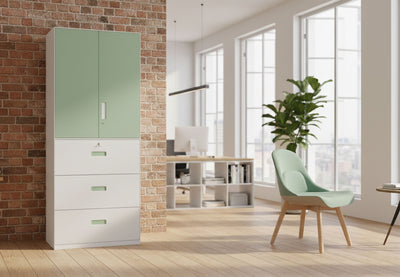 Moda Dual Office Filing Cabinet in a modern office interior with exposed brick walls, wooden flooring, high ceilings, tall windows, and indoor greenery. The tall powder-coated steel cabinet with ABS handles and secure front lock is shown in a styled workspace for e-commerce presentation.