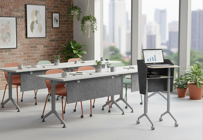 Swish Adaptive Presentation Podium in white oak and black shown in an interior image, positioned at the front of a classroom-style space with four tables facing it, surrounded by high ceilings, large windows and exposed brick walls; modern mobile presentation podium designed for professional training, teaching and collaborative office environments.